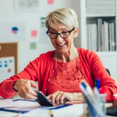 Uśmiechnięta starsza pani w okularach i czerwonym sweterku sprawdza coś na telefonie, zastanawiając się, ile można zarabiać na emeryturze.