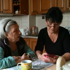 Dwie kobiety przy stole w kuchni dyskutują. Jedna z nich sprawdza telefon, być może szukając informacji, ile kosztuje godzina pracy opiekunki seniora.